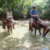 Two people riding horses through a stream in the jungle, navigating the lush and vibrant wilderness with Salto La Jalda (Hiking and Swimming).